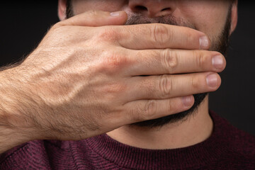 Close up portrait of scared young man covering with hand mouth isolated on black background. Human emotions face expression