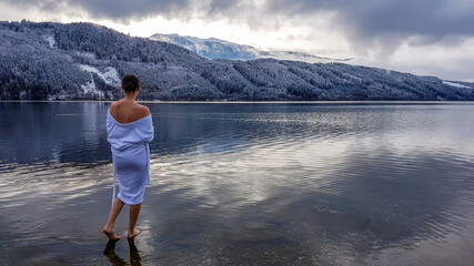 A girl in a white bathrobe walking in a shallow water of a lake. Half of the back of a girl is...