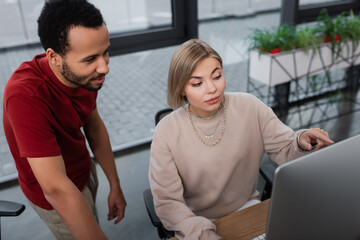 high angle view of blonde manager pointing at computer monitor near african american colleague.