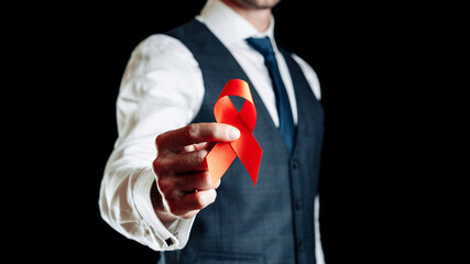Fototapeta premium Aids awareness. Red ribbon symbol in hiv world day in man hand isolated on dark background. Awareness aids and cancer. Healthcare and medical concept.