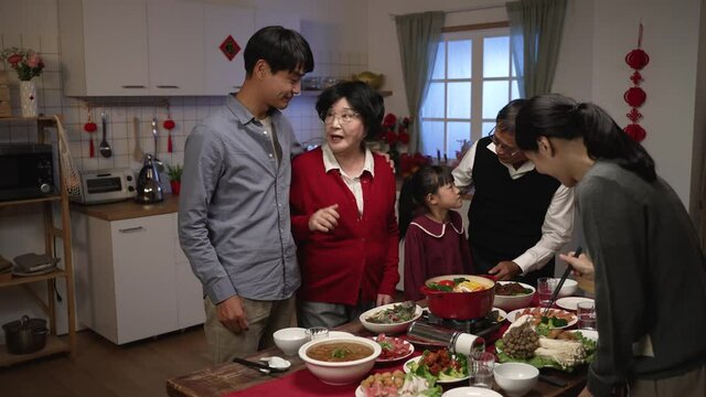 asian senior mother wearing red clothes talking to son while daughter in law preparing hot pot at table. getting ready for chinese lunar new year reunion dinner. chinese text translation: fortune