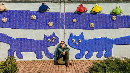 A young man wearing a green leather jacket sits under a mosaic of two cats. Solo traveling man...