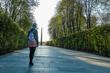 A girl wearing a beanie and a small backpack walks through the alley in a park in Kiev, Ukraine. Both sides of the fence are trimmed nicely. Trees growing on both sides of an alley.