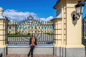 A girl with long hair and in a shot dress sits on a gate leading to a palace. View on the alace is disturbed by the fence. CLear and blue sky. Girl is enjoying her time.