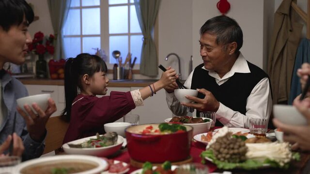 happy asian grandfather talking with hand gestures as his polite granddaughter putting food into his bowl at dinner table on chinese lunar new year's eve at home