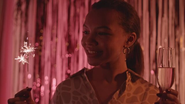 Slowmo close up portrait of young beautiful biracial woman posing with sparkler and champagne glass against red foil fringe curtain