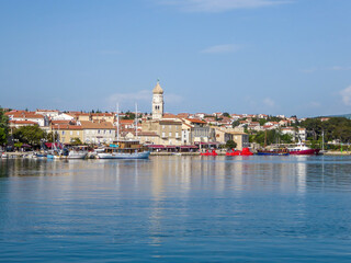 Shore line of city center of Krk, Croatia and the port. In the back there is a church tower. Lots of yachts and submarines parked in the port. There is a forest in the back. Gloomy weather