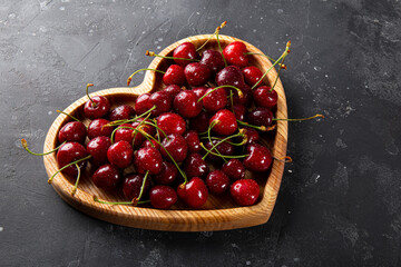 Ripe cherries and strawberries on a wooden plate in the shape of a heart and dark gray background, Valentines card with fruit