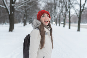 Portrait of young cheerful woman in red knitted hat walking through winter park.