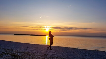 Fototapeta na wymiar A young woman standing on the beach and watching sunset. The beach is pebbly and overgrown with grass. Sun reflects itself in the calm water of the lake. Sunrise above the lake. Freedom and happiness