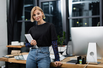 cheerful blonde woman in glasses holding digital tablet near desk in office.