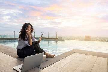Thoughtful concerned asian woman working on laptop and using tablet looking away thinking solving problem in evening at pool on skyscraper, serious woman search for inspiration. Copy space