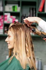 Fototapeta premium Creating curls with curling irons. Hairdresser makes a hairstyle for a young woman with long red hair in a beauty salon