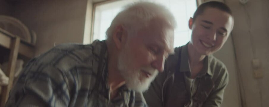 Experienced senior potter shaping clay on wheel and explaining how to make ceramic pot to young woman in workshop