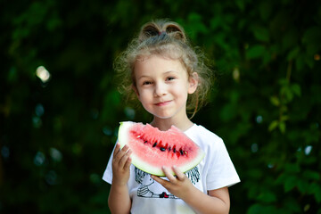 A cute smiling girl eats a red watermelon. Background of green leaves. Nature. Food. Summer.