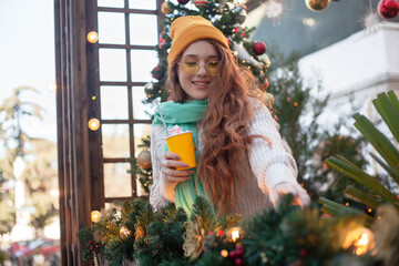 X mas time, red hair girl walking in park on a cold day, enjoying cup of hot chocolate or cocoa, wearing warm in head wear, yellow hat, knitted sweater against the decorated lights