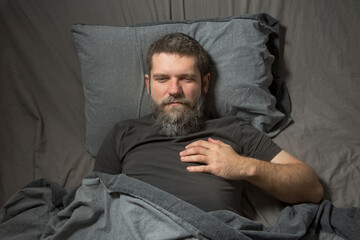 a gray-haired young man with a beard is lying in bed and holding his chest in the heart area with his hand