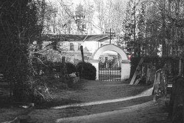 pathway out of cemetery. Cemetery gate with arc and catholic cross, give way road sign and old abanoned building outside graveyard. Black and white with red color left