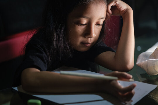 Little Asian girl with long hair is writing in pencil on a table at home, on a dark background. Kid doing homework.