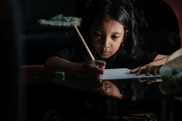 Little Asian girl with long hair is writing in pencil on a table at home, on a dark background. Kid doing homework.