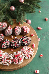 Wooden board with tasty candy cane cookies, fir branch and Christmas balls on green background, closeup