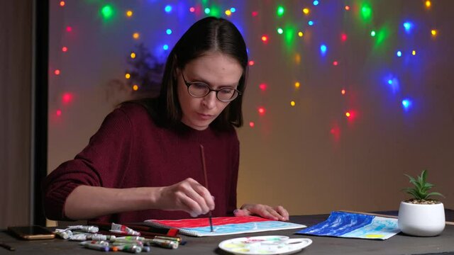 Young talented female artist drawing colorful drawing with the help of paints and brushes, sitting on the table in the evening at home on background of christmas decorations garlands. 