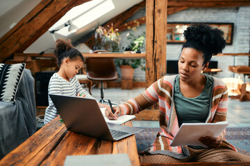 Black working mother takes notes while analysing business reports at home.