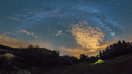 Panorama of Milky Way bow with illuminated tent besides a river and forest, Croatia