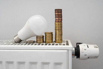 Energy crisis. White LED light bulb on a radiator with money coin stack. Growing electric and gas bill. Central heating.