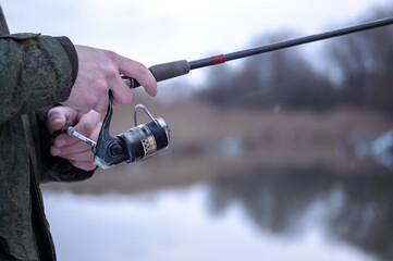 The frozen hands of a male angler close-up to hold the spinning in a blurry motion wind the fishing line on a reel against the background of a winter river. Selective focus on the hand. Background