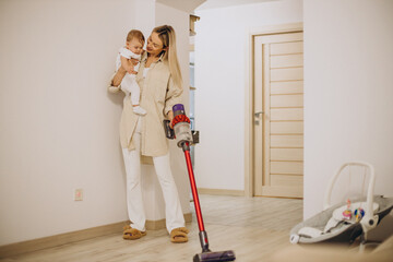 Mother vacuuming with baby daughter at home with the help of rechargeable vacuum cleaner