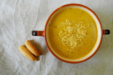 a soup of zucchini, onions, potatoes and carrots, sprinkled with grated hard cheese, in a brown plate with handles stands on a gray tablecloth with two small Italian loaves. home healthy kitchen. home