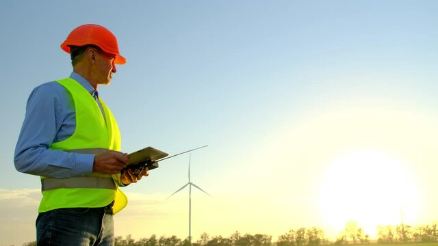 Windmills Generate Clean Power Against Bright Sun. Professional Engineer Stands In Field Checking Data In Tablet Near Rotating Turbines