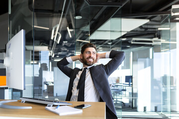 Happy and dreamy businessman, resting at work with his hands behind his head, working in a modern office at the computer