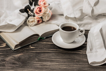 Morning coffee. A cup of coffee with a saucer on a wooden table, an open book and a white shirt on the background of a bouquet of flowers. Still life concept. Cozy morning.