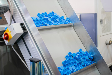 Many blue plastic bottle caps moving on conveyor belt of automatic compression molding machine at factory, exhibition: production line. Manufacturing, recycling, industry, technology equipment concept - obrazy, fototapety, plakaty
