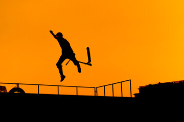Unrecognizable teenage boy silhouette showing high jump tricks on scooter against orange sunsetwarm...