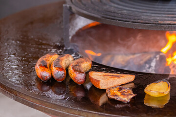 Process of grilling fresh meat sausages, toast bread slice, pepper on round brazier with hot flame at summer local food market: close up. Outdoor cooking, barbecue, gastronomy, street food concept