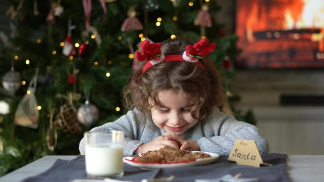 Little funny girl eats a cookie that she has prepared for Santa Claus. Little girl waiting for Santa Claus with cookies and a glass of milk on Christmas eve