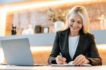 Happy smart caucasian businesswoman, tom manager, ceo, sitting at a table in a modern office, using a laptop, pondering business strategies and financial opportunities, taking notes, smiling