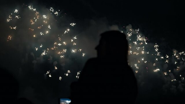 The silhouette of a woman who shoots a huge multi-colored fireworks on a black sky on her smartphone at night. 