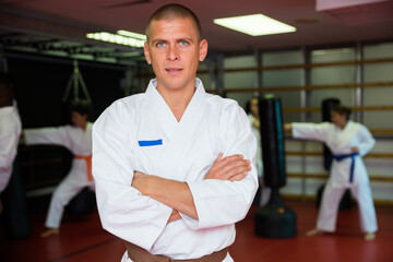 Caucasian man karate trainer standing with crossed hands in gym during group training. Women exercising in background.