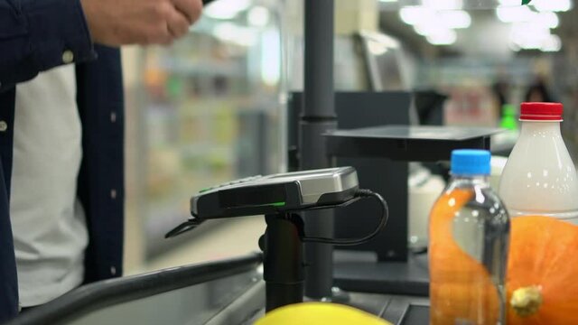 Client paying for purchases by card and taking bill, standing at checkout of store spbd. Closeup view of young male buyer pays for organic food and uses payment service, cashier makes sale and sits at