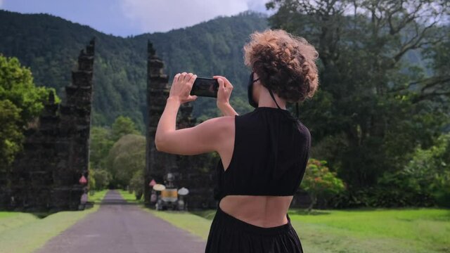 Happy tourist blogger girl in face mask tacking photos on Traditional Balinese Hindu gate Candi Bentar, Bedugul, Indonesia. Exciting vacation trip to Bali