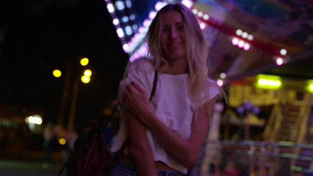Young blonde woman models for camera at showground carnival at night. Handheld shot.