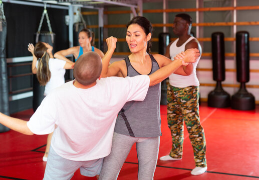 Asian woman training elbow strike with caucasian man in gym during self-defence training. African-american man trainer standing nearby and observing.