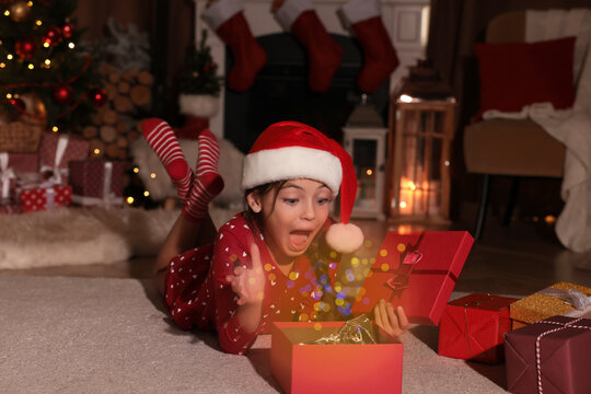 Surprised child in Santa hat opening Christmas gift on floor at home