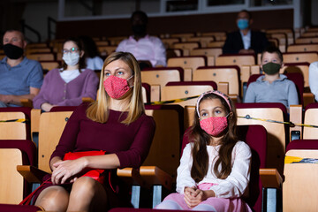 Young woman with preteen daughter wearing protective masks watching theatrical performance. Concept of precautions during coronavirus pandemic