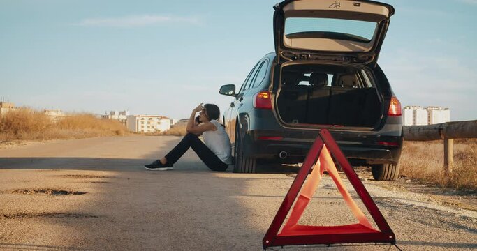 Young woman sitting a side of breakdown vehicle with flat tire using smartphone, car blinking emergency lights and warning triangle on country road during travel to home. Focus on triangle. 