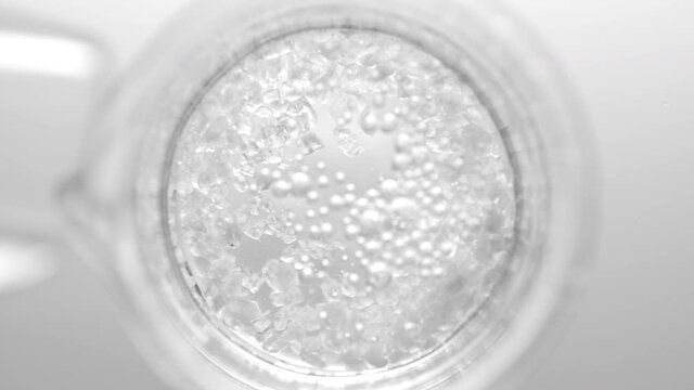 Top view macro shot of crystals fall down from test tube into clear fluid in flask creating different sized bursting bubbles on light grey background | Mineral skincare cosmetics formulating concept